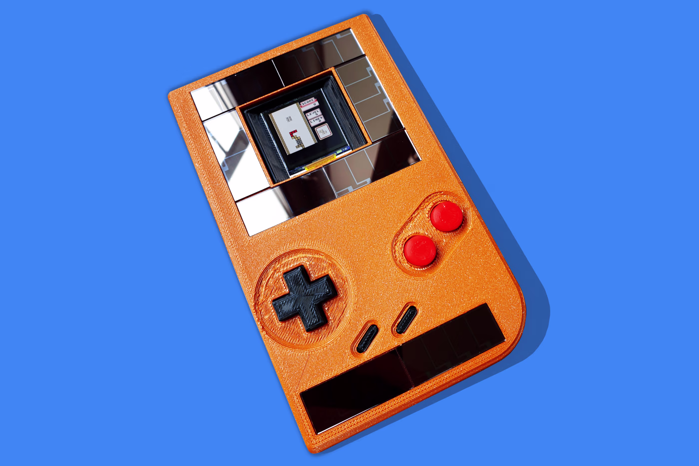 The battery-free Game Boy: an orange 3D-printed handheld with a solar panel, mirrored e-paper-style display, and directional pad, photographed for the Wall Street Journal.